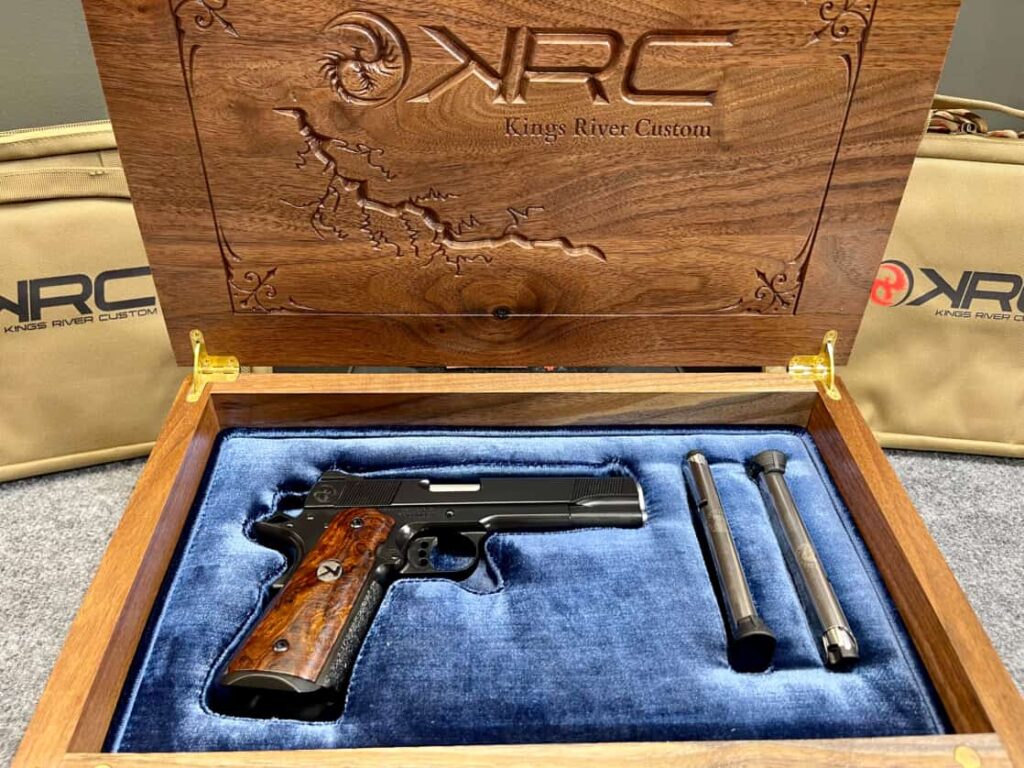 A wooden box containing a custom 1911 pistol on a blue padded insert with two spare magazines.