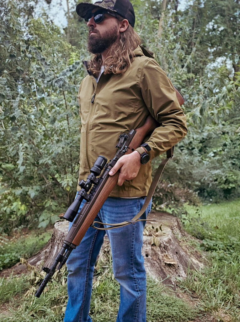 Man standing in raincoat with an M1A Scout Squad under his arm.