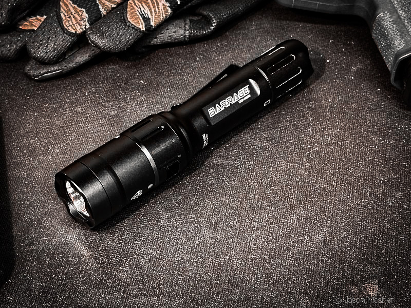 A black flashlight on a table that is turned on. The words "Barrage" are printed across the side of the light. 