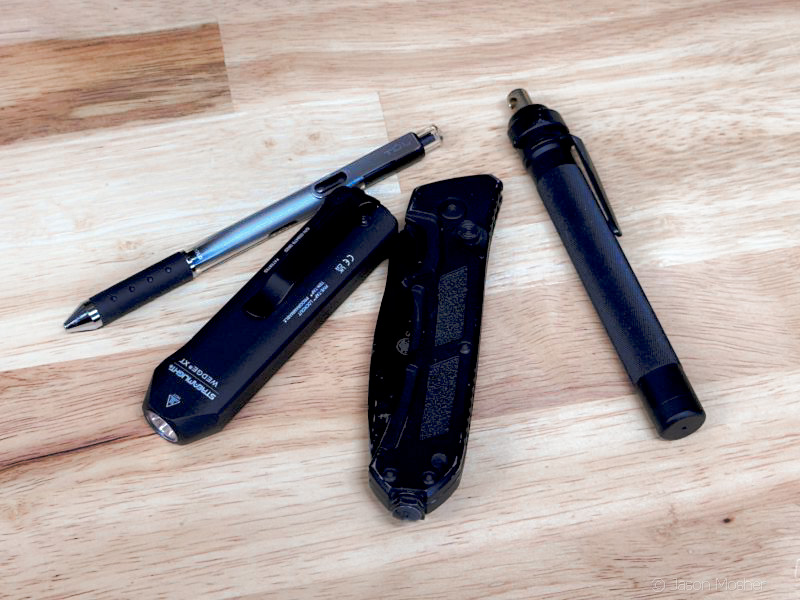 A photo of a pen, flashlight, black pocket knife, and a small rube of pepper spray on a wood counter. 