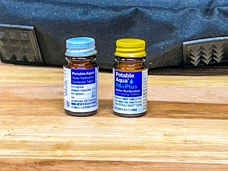 Two small bottles, one with white lid and one with a yellow lid of Potable Aqua tablets on a wood table.