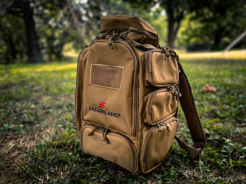 A brown backpack sitting on the ground with green grass around it. The name "Safariland" is visible in red stitching across the front of the bag.