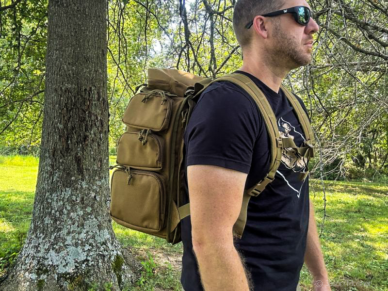 A person wearing a black shirt and brown backpack, walking. Trees and green grass can be seen in the background.