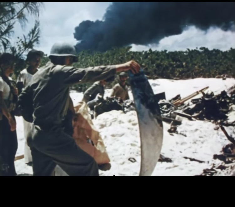 In this still from the 1942 U.S. Navy film” The Battle of Midway”, shot by John Ford, soldiers and civilians inspect the wreckage of a plane while black smoke billows in the distance. (US. Navy. Public Domain)