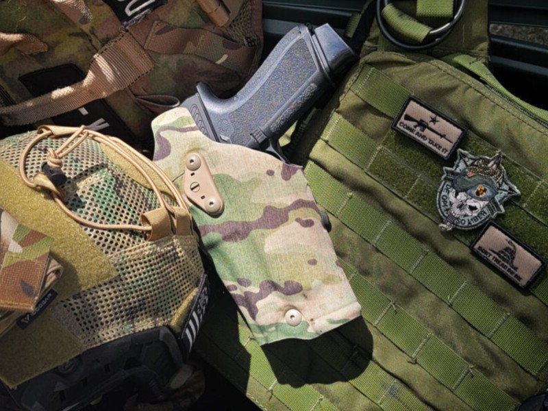 A camo holster next to an army helmet and camo vest. A black gun is in the camo holster. 