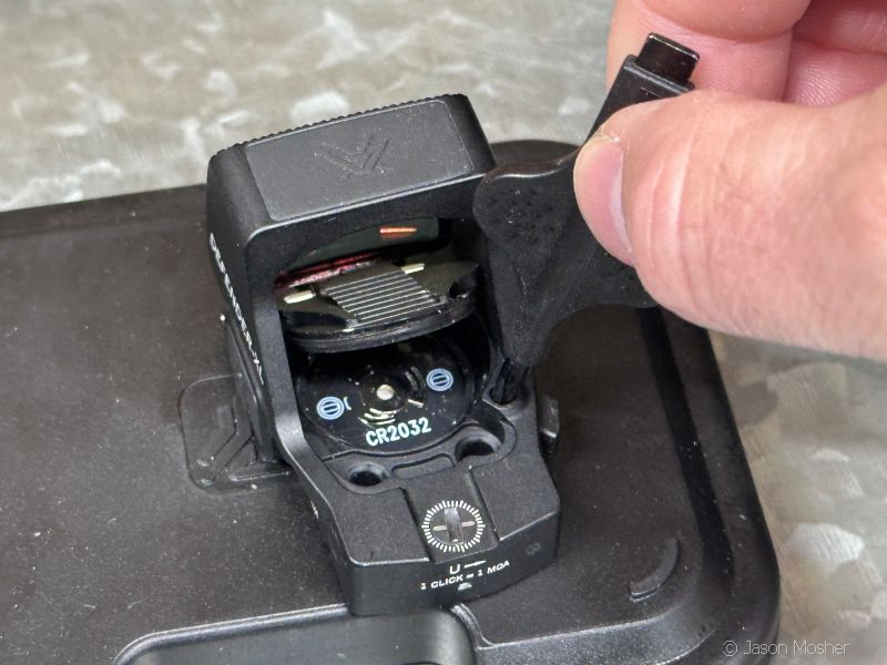 close up of hand using a small tool to press a button on the back of the Vortex Defender XL red dot to open the battery door. 
