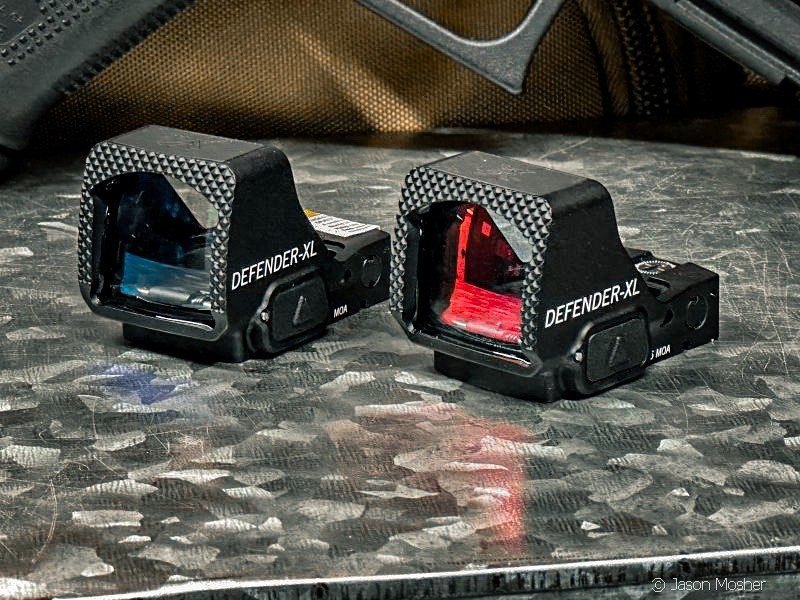 Two Vortex handgun optics side by side on a metal surface. One has a green tint to the glass and the other has a red tint.