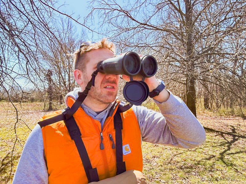 A person standing in the woods wearing an orange vest, looking through a pair of binoculars. 