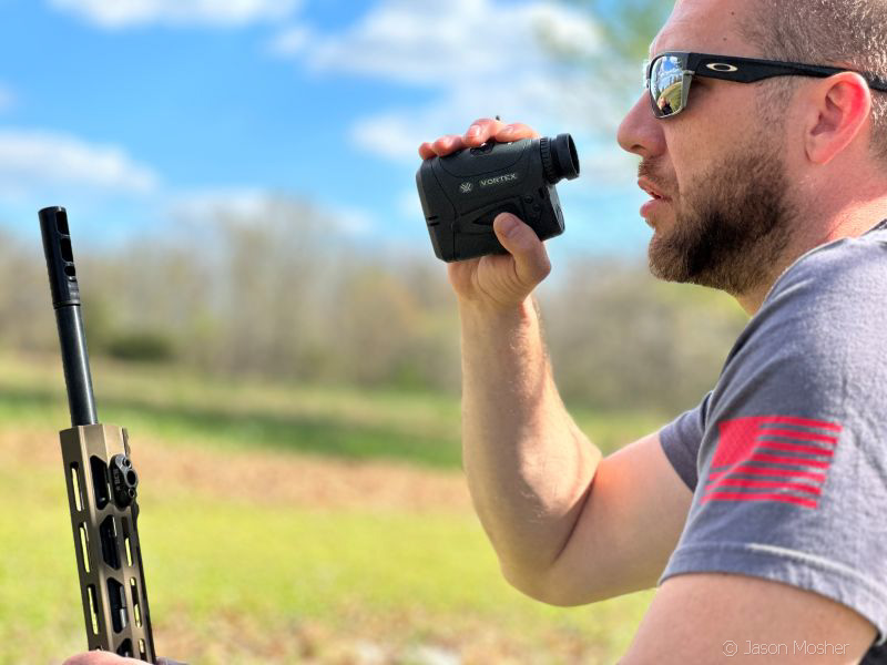 A person kneeling down, holding onto the side of their rifle with one hand and looking through a range finder with the other. A blue sky is in the background and the person is wearing sunglasses and a grey t-shirt. 