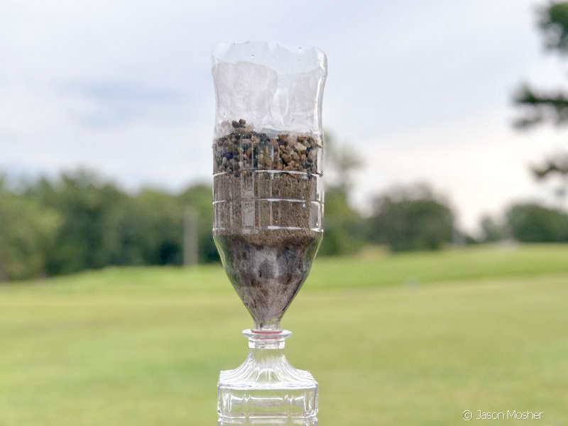 homemade water filter: A pop bottle turned upside down, the the bottom cut-off and now the top. A black area at the bottom is activated charcoal, follwed by some sand and then gravel. The green grass is visible in the background.