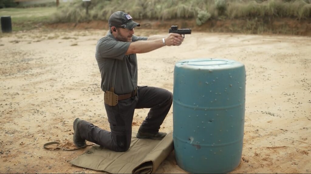 Rick Hogg of War HOGG Tactical is demonstrating shooting a pistol from a kneeling position behind a pickle barrel for simulated cover
