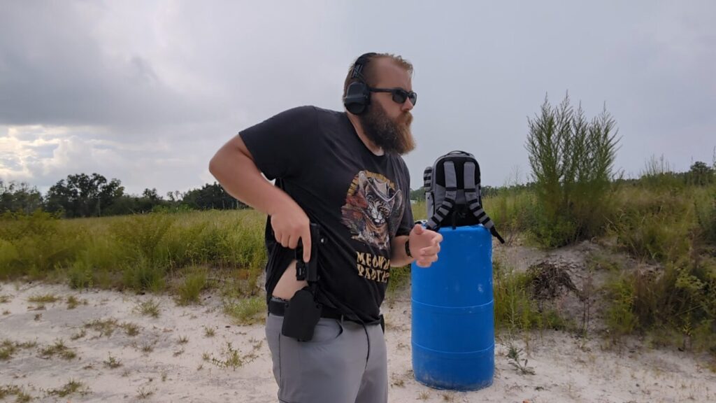 A man wearing a "Meowdy Partner" t-shirt and sunglasses at a shooting range, drawing a Ruger RXM pistol from a holster at his waist.