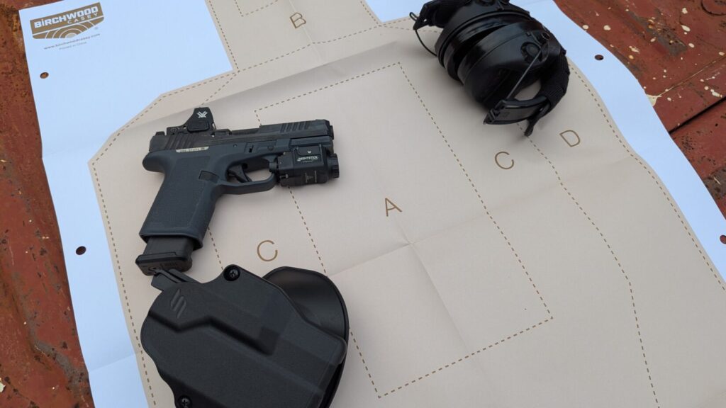 A black Ruger RXM handgun with a Vortex Defender-ST red dot sight and a weaponlight, a black Safariland paddle holster, and a pair of black electronic ear protection headphones are shown on a USPSA target