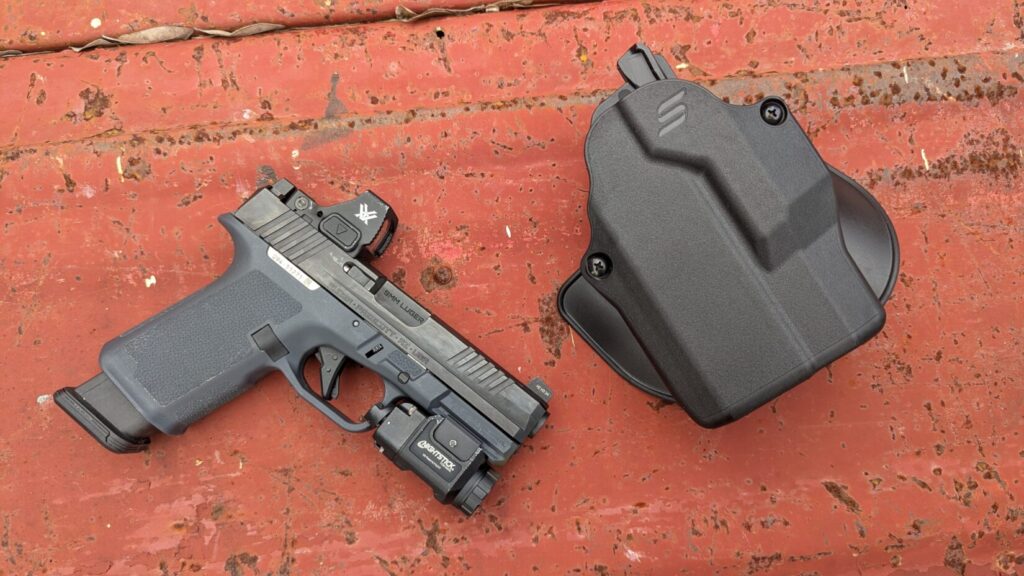 A black Ruger RXM handgun with a Vortex Defender-ST red dot sight and a weaponlight is next to a black Safariland holster on a rusty red surface.

