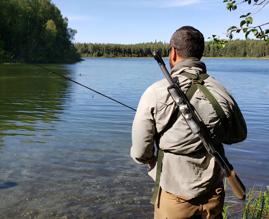 A man fishing at a lake with a rifle slung over his back.