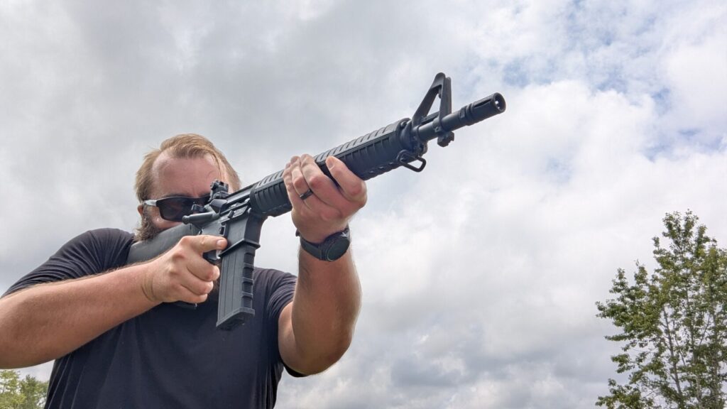 Man wearing sunglasses, aiming a black dissipator rifle in a field with a cloudy sky.