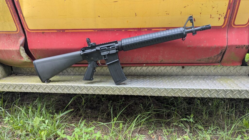 A black dissipator rifle with a fixed stock, leaning against a red and yellow truck.
