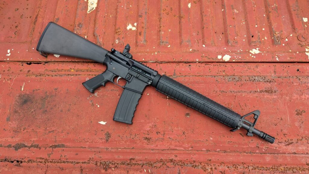 A black dissipator rifle with a fixed stock and a magazine inserted, lying on a red, textured metal surface.