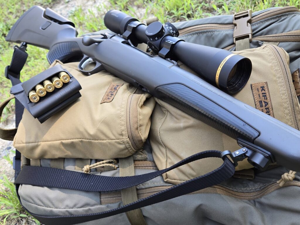 A bolt action rifle with a magnified optic resting on top of a tan and ranger green backpack with spare ammunition nearby.