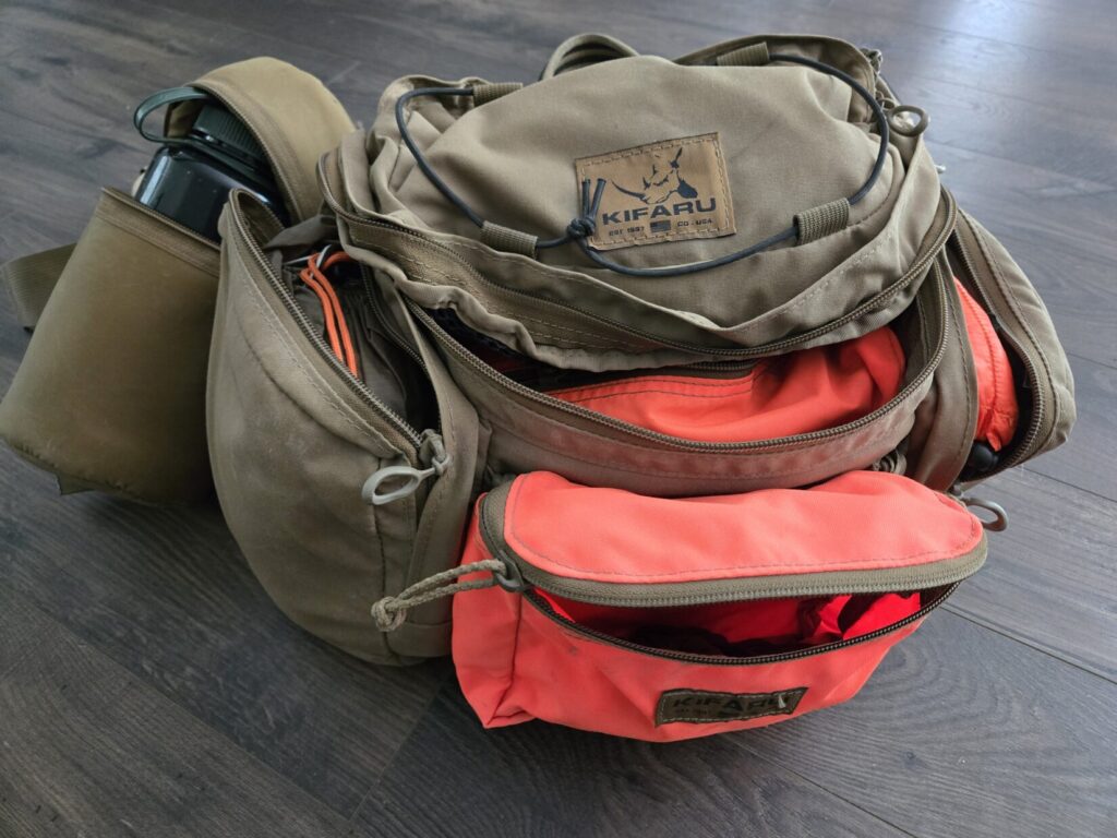 A tan nad blaze orange lumbar pack from Kifaru with zippers open along with a padded water bottle pouch.