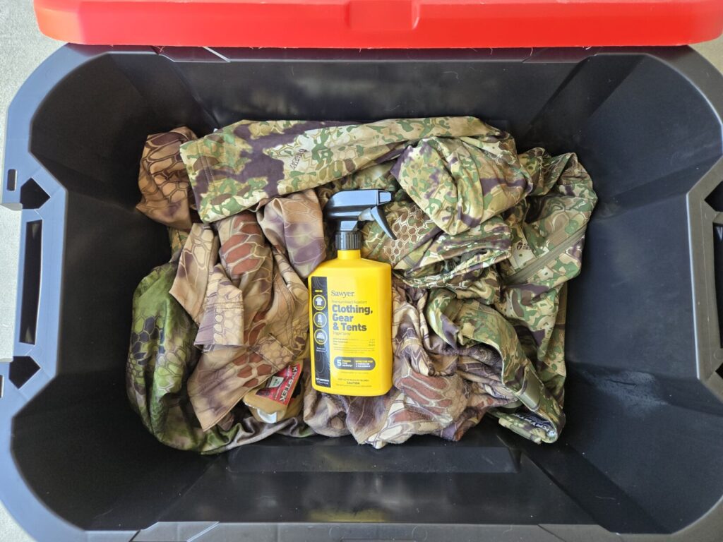 Kryptek camouflage clothing inside a plastic bin with Sawyer Permethrin on top of it