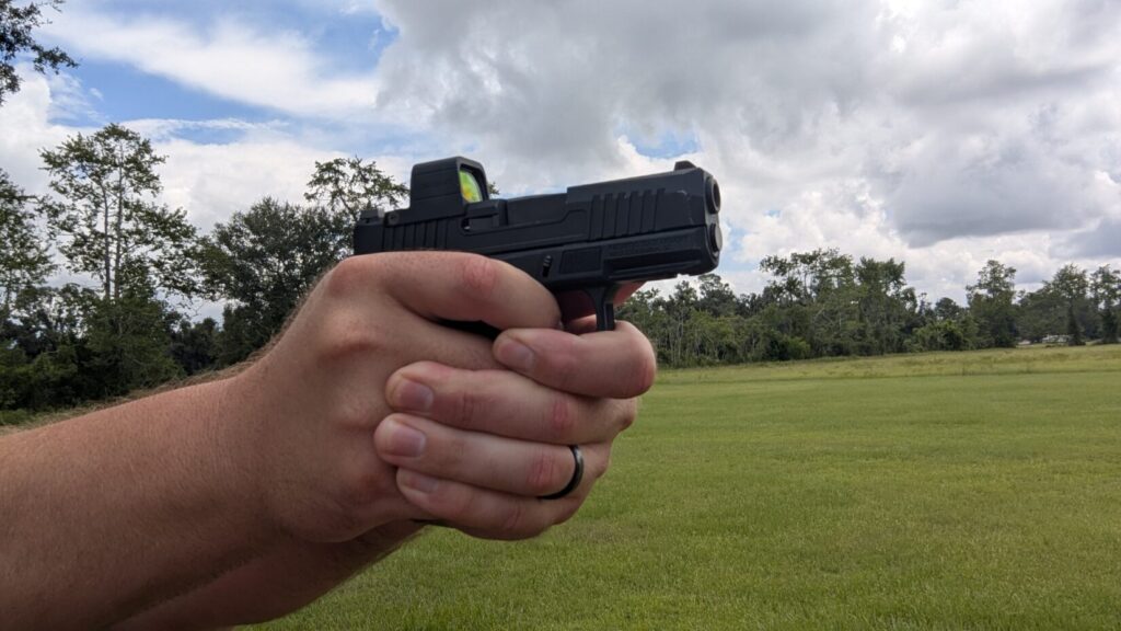 A person's hands holding a PSA  pistol with an optic, aiming to the right against a backdrop of a cloudy sky and a green field.