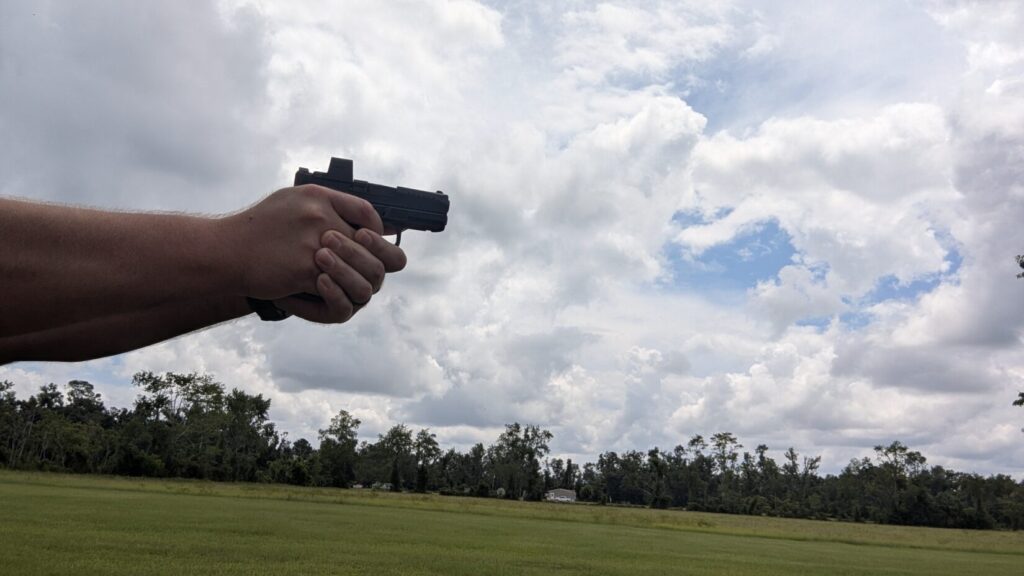 A person's hands holding a Palmetto State Armory (PSA) Micro Dagger pistol with an optic, aiming forward in a grassy field under a cloudy sky.
