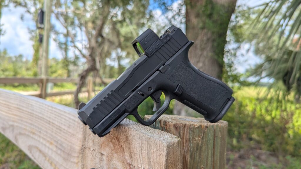 A PSA pistol with a red dot optic mounted on a wooden fence post in a rural outdoor setting.