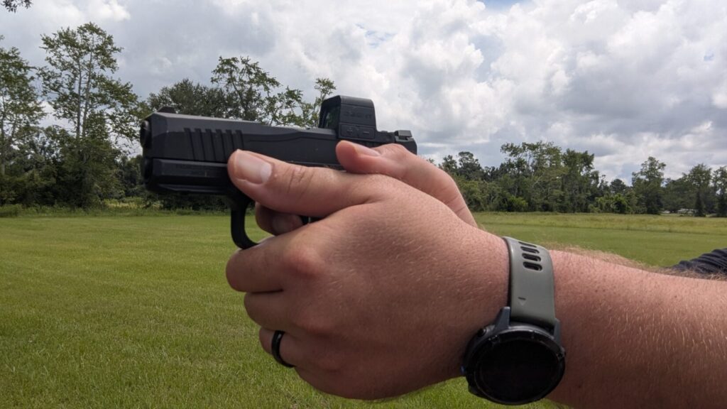 Person aiming a black handgun with an Osight S red dot sight in a field.