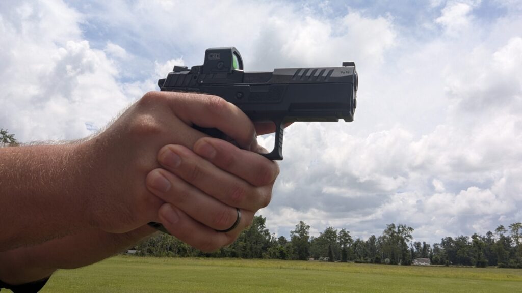 A person's hands holding a black handgun with an EDC Enclosed red dot sight, aiming it in a field. The background shows trees and a cloudy sky.