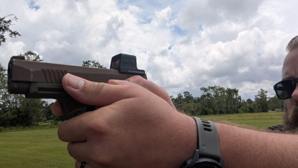 Person's hands holding a tan and green handgun with a Holosun EPS Carry red dot sight, aiming it in a field.