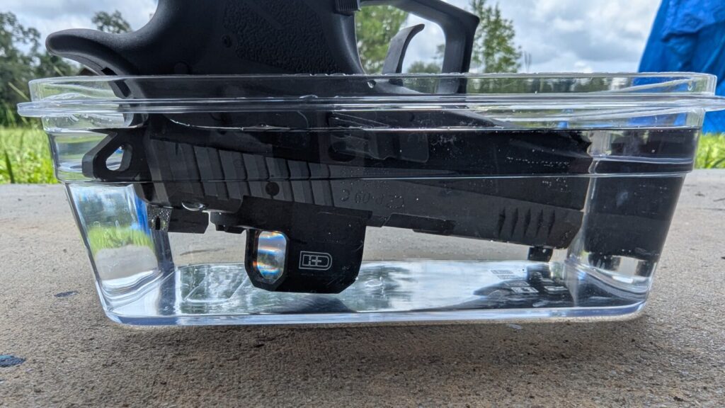 Black handgun with an EDC Enclosed red dot sight submerged in a clear plastic container of water.