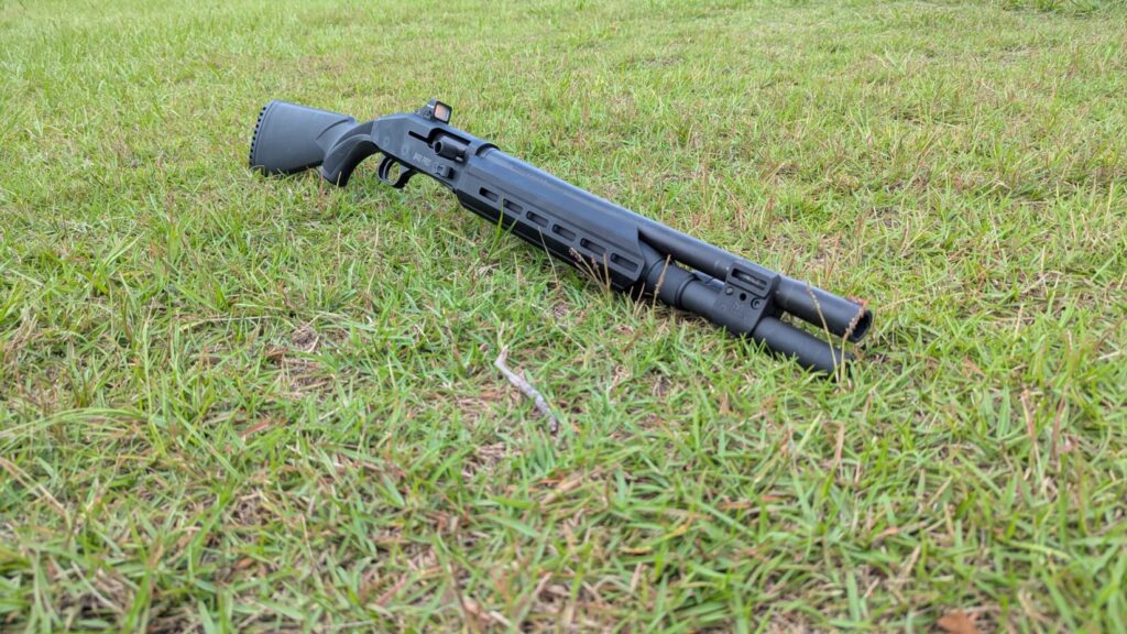 A black Mossberg 940 Pro Tactical shotgun with a Holosun red dot sight, lying on a grassy field.
