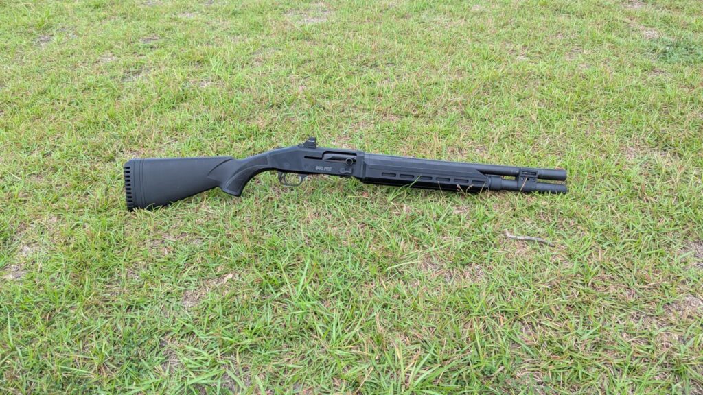 A black Mossberg 940 Pro Tactical shotgun with a Holosun red dot sight, lying on a grassy field.
