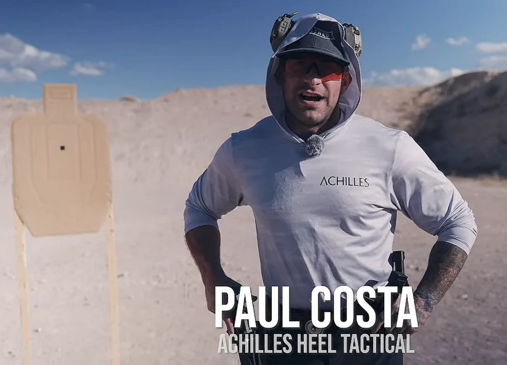 Achilles Heel Tactical's Paul Costa on an outdoor shooting range, wearing ear and eye protection, facing the camera. Cardboard sillhouette is in the background to his right, berm in the distance.