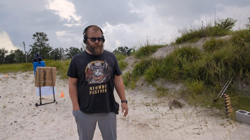 A man with a beard, sunglasses, and ear protection stands at an outdoor shooting range.