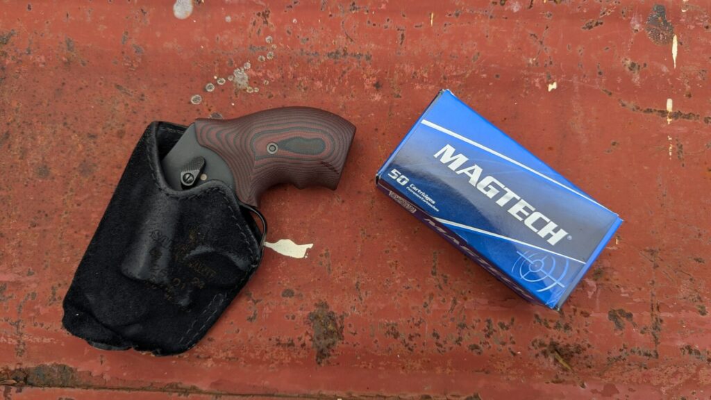 A black Smith & Wesson 432 UC revolver with a wooden grip lies next to a black suede-like holster on a rusty red background.
