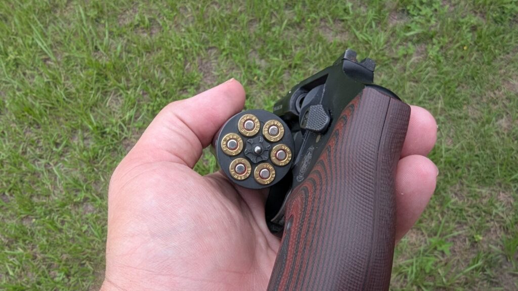 A hand holds a loaded Smith & Wesson 432 UC revolver, showing the 6 rounds in the cylinder against a grassy background.