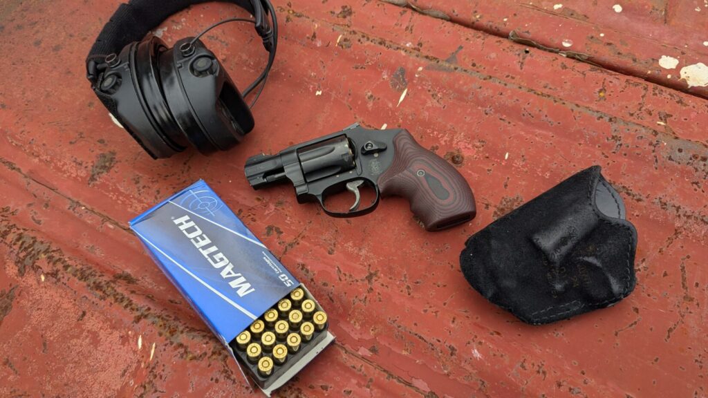 A black Smith & Wesson 432 UC revolver with a brown wooden grip, a black holster, and a blue box of Magtech ammunition are arranged on a rusty red surface. A pair of black electronic hearing protection headphones are also visible.