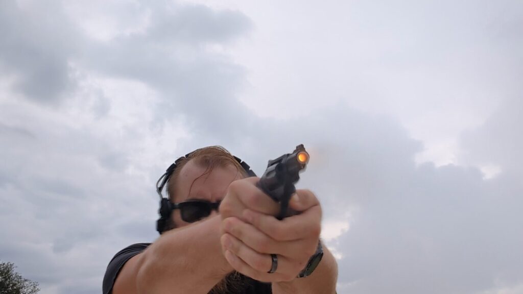 A man in sunglasses and ear protection aims a black Smith & Wesson 432 UC revolver, with a bright muzzle flash visible.