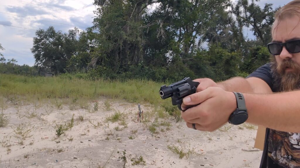 A man in sunglasses and ear protection stands in a sandy, outdoor range, holding a black Smith & Wesson 432 UC revolver with arms extended, aiming at a target.