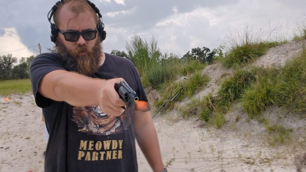  A man in a "Meowdy Partner" t-shirt, sunglasses, and ear protection, holds a black Smith & Wesson 432 UC revolver with a muzzle flash visible, aiming it at the camera.
