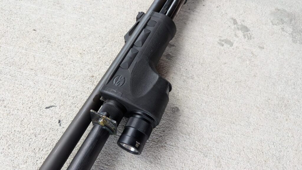 A close-up of a shotgun's forend with a Surefire DSF weaponlight, resting on a wooden surface. The Surefire logo is clearly visible.
