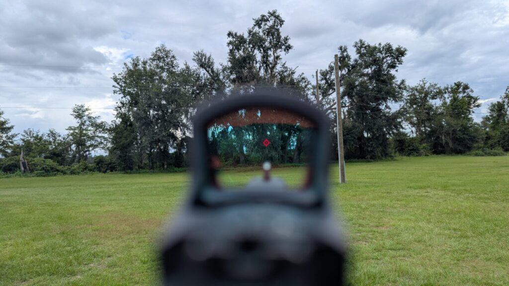 A view through the Swampfox Justice 2 optic's window, showing a red dot aiming at a target in a grassy field.