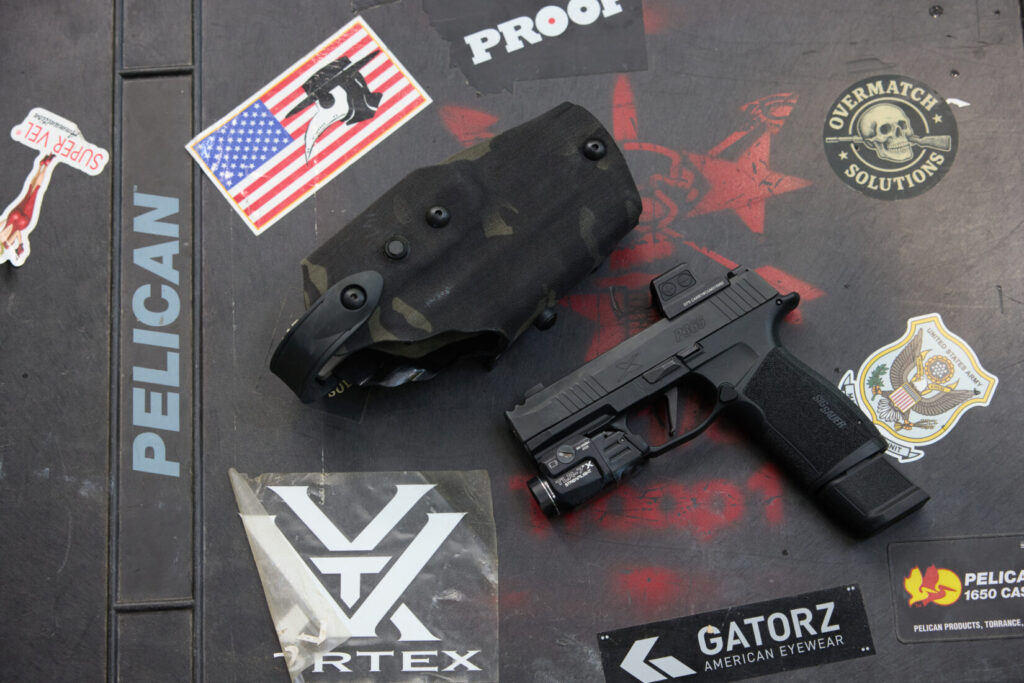 A close-up shot of a black SIG P365 XMACRO handgun with a mounted light and optic, next to a black camouflage Safariland 6360RDS holster,