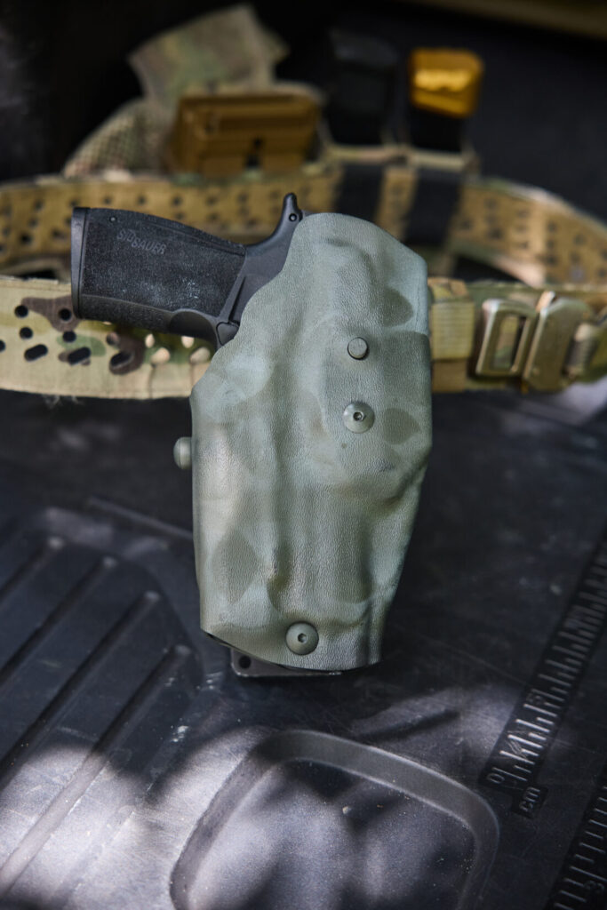 A close-up, angled view of a Sig P365 XMACRO holstered in a Safariland holster, which is attached to a tactical belt.