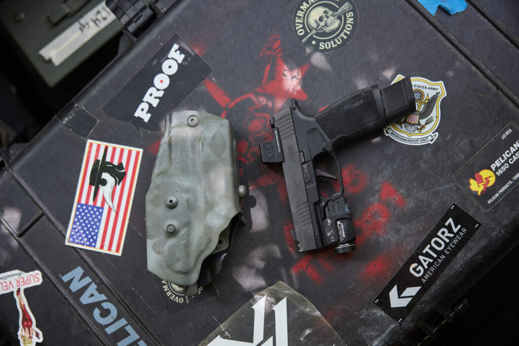 A black handgun with an attached light and optic is positioned next to a greenish-gray holster
