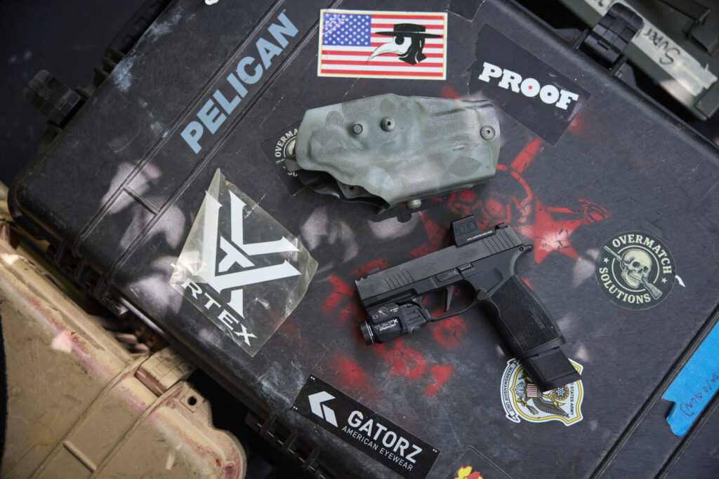 A black handgun with an attached light and optic is positioned next to a greenish-gray spray-painted holster