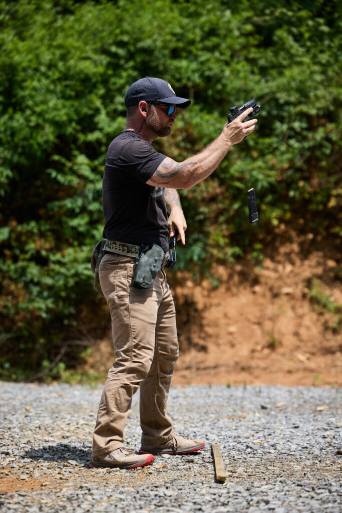 A man at an outdoor shooting range is shown mid-action, ejecting a magazine from his handgun while holding it with one hand.