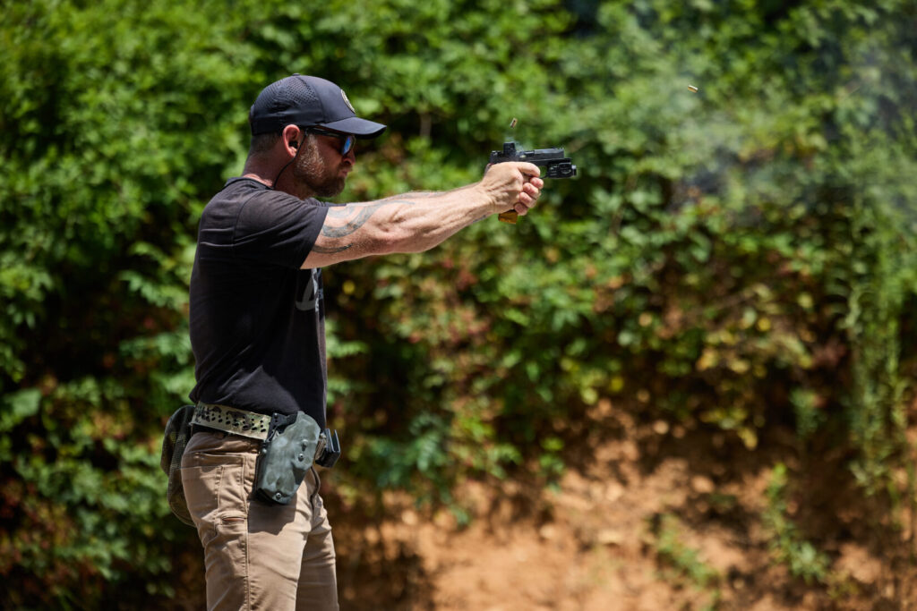 A man shooting a Sig XMACRO at a range, with a small puff of smoke and a spent casing visible near the firearm's ejection port.
